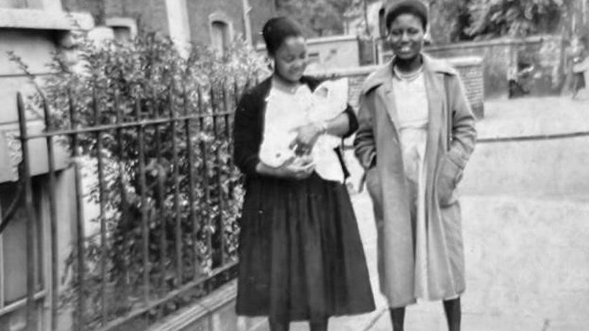Dame Ijeoma as a baby is being carried by her godmother, and her mother is smiling beside her in a black-and-white photo. Both women are wearing long coats that reach their knees and dresses of the same length. They are standing in the street, near the gate of a house.