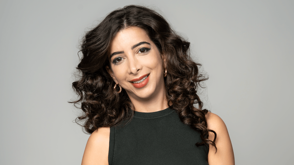 Žena duge tamne, kovrdžave kose, sa zlatnim okruglim minđu&scaron;ama na u&scaron;ima i crvenim karminom, nasmejana gleda u kameru, glave blago nagnute na levo, u tamnoj majici bez rukava