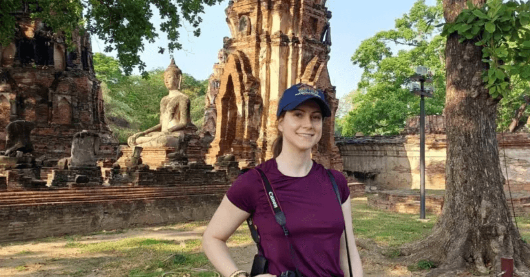 Devojka sa bejzbol kapom u ljubičastoj majici i fotoaparatom oko vrata ispred statue Bude na Tajlandu