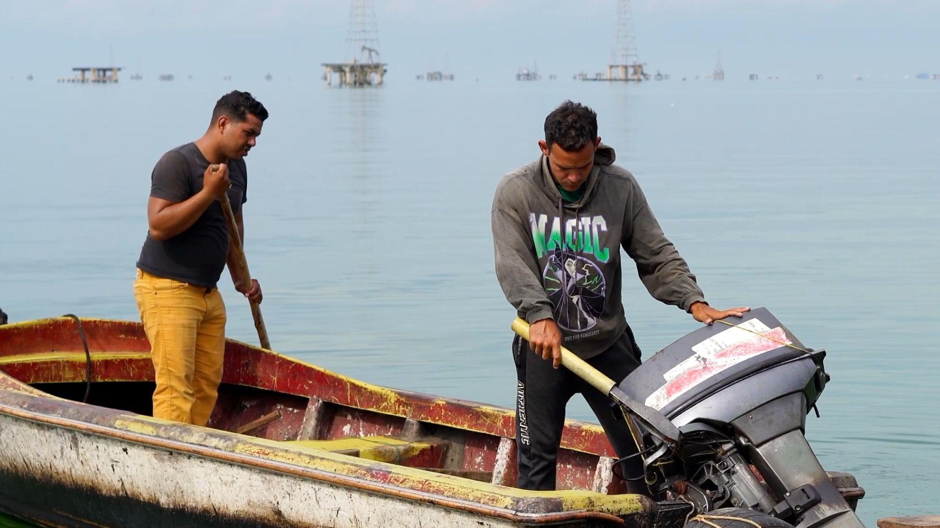Dva mladića stoje u malom otvorenom čamcu na jezeru. Onaj levo, u žutim pantalonama i crnoj majici, drži drveni &scaron;tap. Momak desno, u sivoj dukserici sa reči Magija ispisanom zelenom i belom bojom, drži ruke na sivom vanbrodskom motoru. Naftne platforme izdižu se iz vode u pozadini.