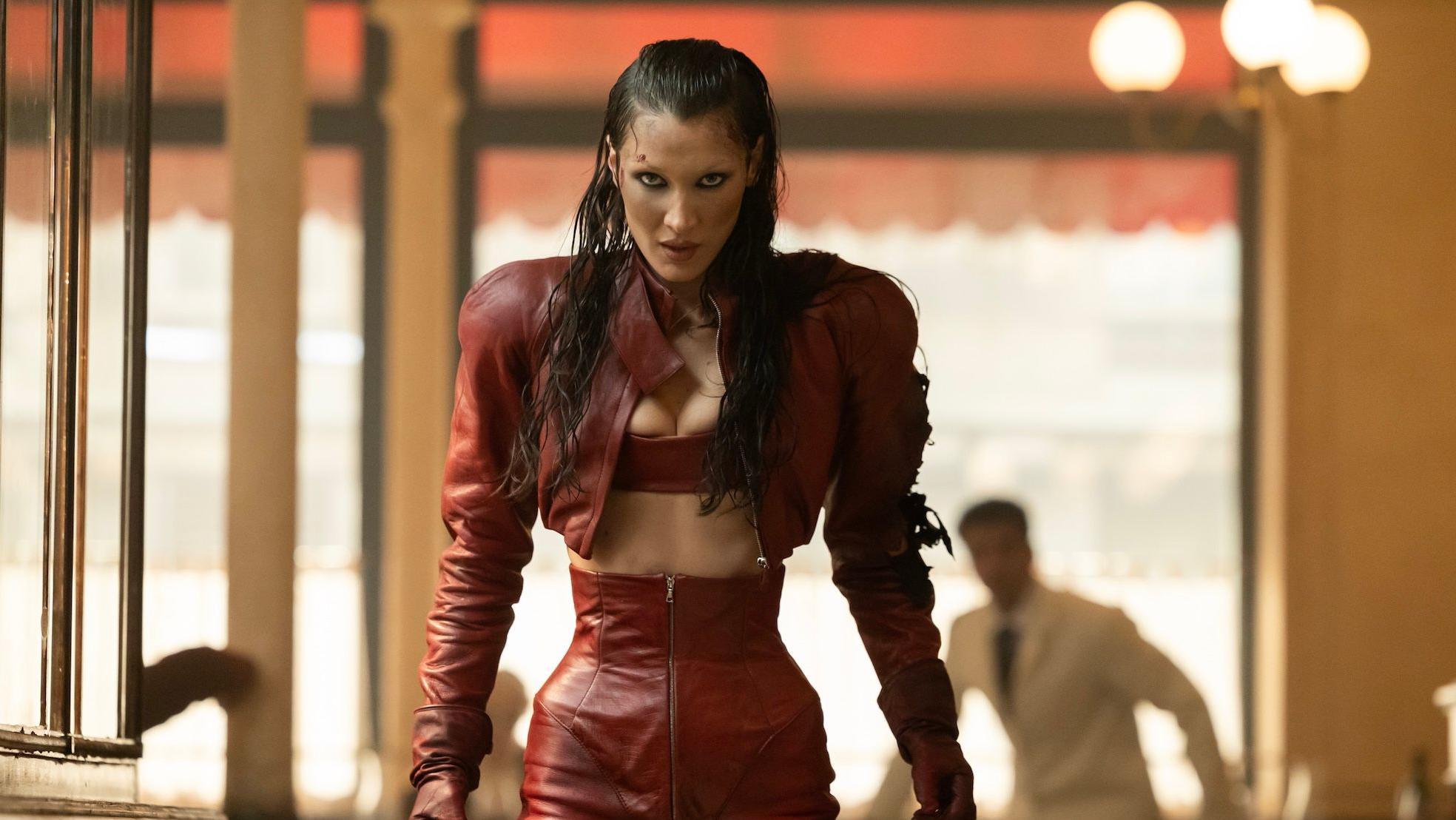 Slika Bele Hadid u crvenom outfitu, sa upla&scaron;enim izrazom lica. Pozadina je zamućena, ali se može videti mu&scaron;karac u beloj ko&scaron;ulji.