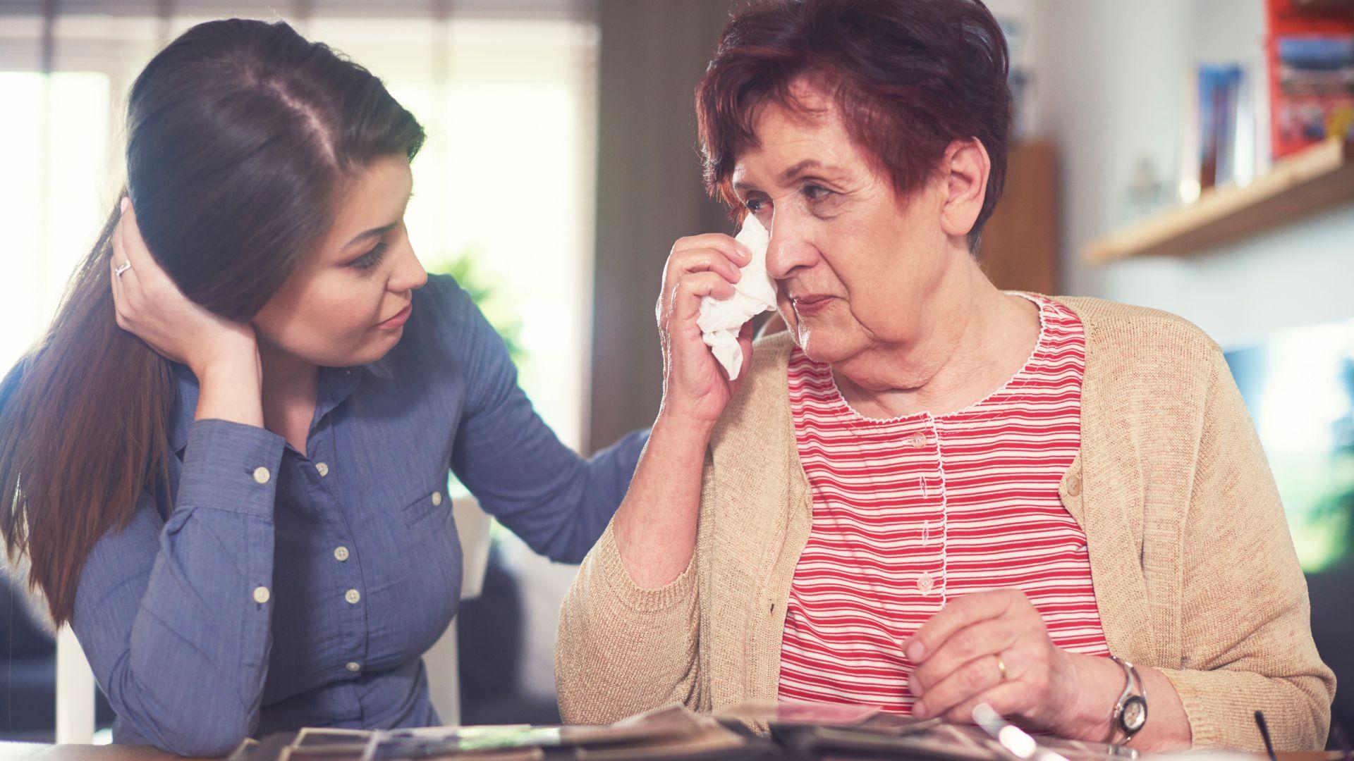 Starija žena drži maramicu na suznom oku dok gleda foto-album; mlađa žena koja sedi pored je te&scaron;i