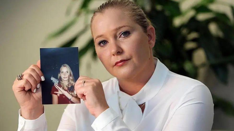 Virginia Giuffre, a woman with blonde hair tied back, wearing a white blouse and holding up a photograph of herself as a teenager.