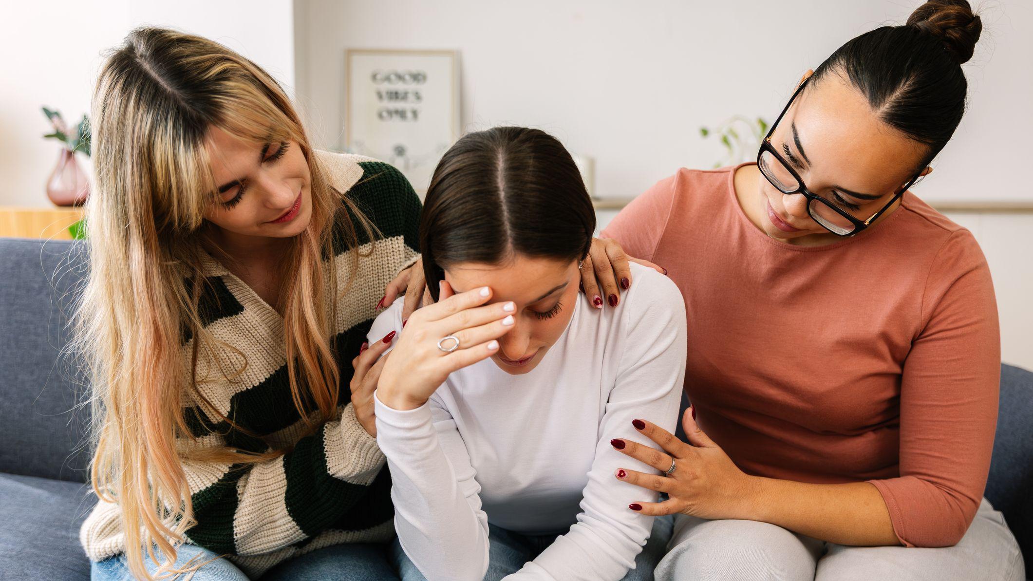 Dve mlade žene te&scaron;e prijateljicu koja sedi između njih na sofi, sa rukom na licu i izgleda uznemireno.